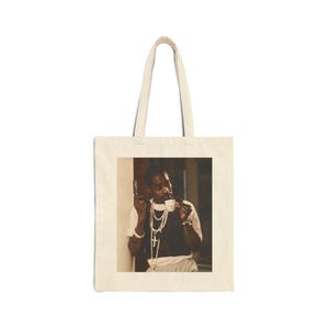 Op de afbeelding: Crèmekleurige draagtas met een sepia-afbeelding van een persoon die een pistool en een kopje thee vasthoudt. De persoon draagt een wit overhemd, een zwart vest en kettingen. De tas heeft een rechthoekige afbeelding en twee handvatten.