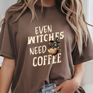 Op de afbeelding: Bruin t-shirt met de crèmekleurige tekst "EVEN WITCHES NEED COFFEE". De afbeelding toont een heks die in een koffiekopje roert. Het t-shirt heeft een casual stijl.