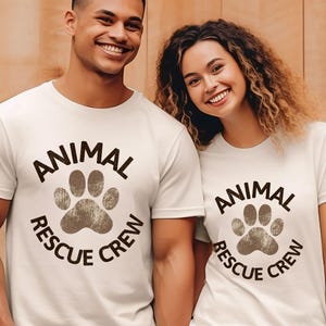 May include: Two people wearing white t-shirts with the text "ANIMAL RESCUE CREW" in a circular design with a paw print. The shirts are a light color, and the text is dark brown.