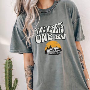 Pu&ograve; includere: T-shirt grigio con la scritta "TWO HEARTS ONE RV" in un font retr&ograve;. Sotto, una grafica di un camper davanti a un sole giallo e alberi. La maglietta ha un aspetto vintage e lavato.