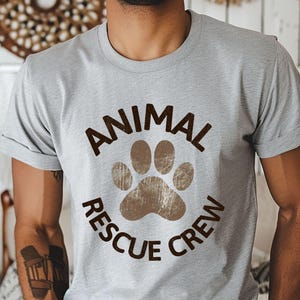 May include: A light gray t-shirt with the words "ANIMAL RESCUE CREW" in brown, encircling a brown paw print. The shirt is short-sleeved and has a crew neck.