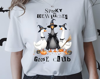 May include: A white short-sleeve t-shirt featuring a graphic of three geese wearing witch hats. The centre goose wears a black cape. The text reads SPOOKY BEWITCHED GOOSE CLUB. Small pumpkins and a broom are included in the colourful illustration.