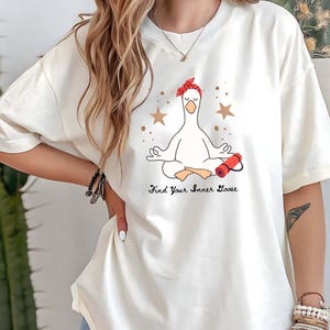 May include: Cream-coloured t-shirt featuring a meditating goose wearing a red bandana and holding a red yoga mat. The text "Find Your Inner Goose" is printed below the goose. The design includes brown stars.