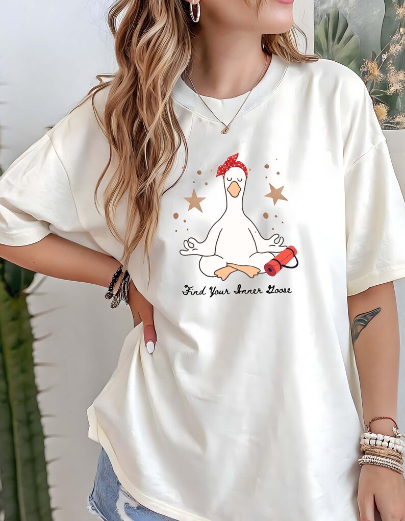 May include: Cream-coloured t-shirt featuring a meditating goose wearing a red bandana and holding a red yoga mat. The text "Find Your Inner Goose" is printed below the goose. The design includes brown stars.