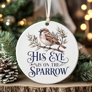 May include: A round white ceramic ornament hanging from a gold ribbon. The ornament features a brown sparrow perched on a branch with green leaves and red berries. The text "His Eye Is On The Sparrow" is printed in dark blue script and serif font.