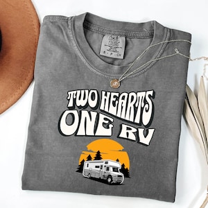 Pu&ograve; includere: T-shirt grigio Comfort Colors con la scritta "TWO HEARTS ONE RV" in un font retr&ograve;. Sotto, un'illustrazione di un camper davanti a un tramonto. Una collana d'argento con un ciondolo &egrave; sulla maglietta.