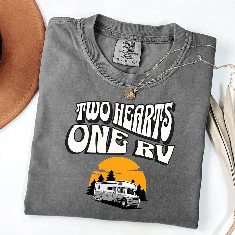 Pu&ograve; includere: T-shirt grigio Comfort Colors con la scritta "TWO HEARTS ONE RV" in un font retr&ograve;. Sotto, un'illustrazione di un camper davanti a un tramonto. Una collana d'argento con un ciondolo &egrave; sulla maglietta.