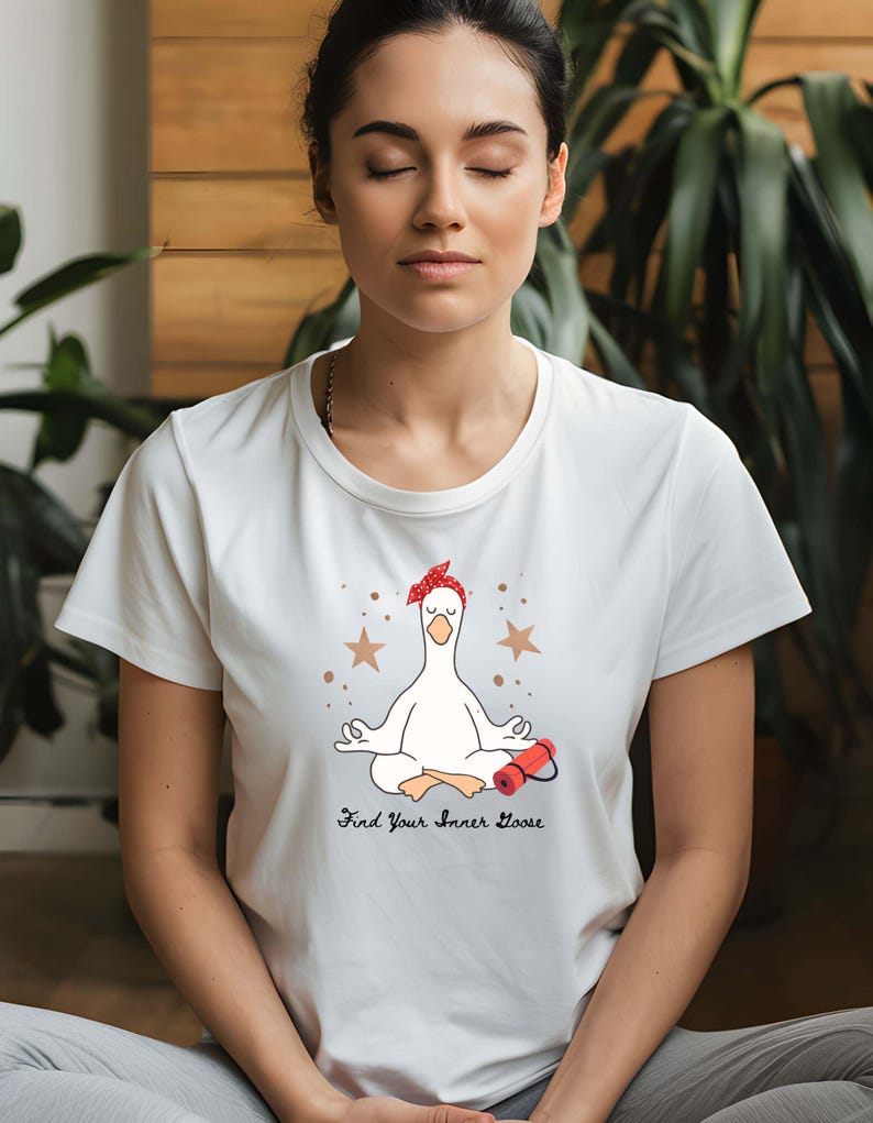 May include: White t-shirt with a cartoon goose in a yoga pose, wearing a red bandana. The text "Find Your Inner Goose" is below the goose. Two gold stars are above the goose. The model is wearing grey trousers.