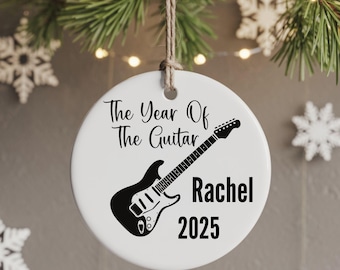 Może przedstawiać: Okrągła, biała ceramiczna ozdoba świąteczna z czarną sylwetką gitary i napisem "The Year Of The Guitar Rachel 2025". Ozdoba zawieszona jest na sznurku, na tle świątecznej zieleni i płatków śniegu.