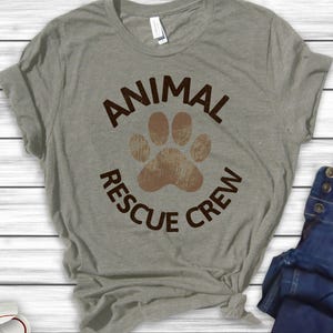 May include: A heather gray t-shirt with the words "ANIMAL RESCUE CREW" in brown, surrounding a brown paw print. The shirt is made of a soft material and is knotted at the bottom.