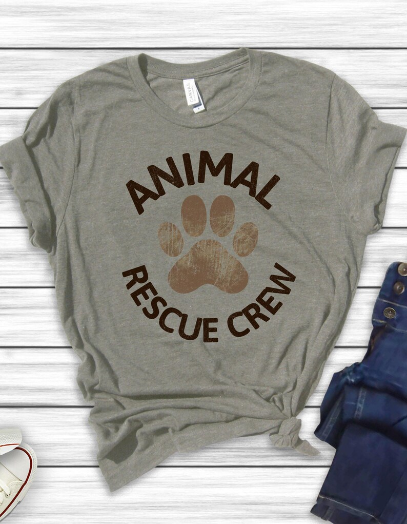 May include: A heather gray t-shirt with the words "ANIMAL RESCUE CREW" in brown, surrounding a brown paw print. The shirt is made of a soft material and is knotted at the bottom.