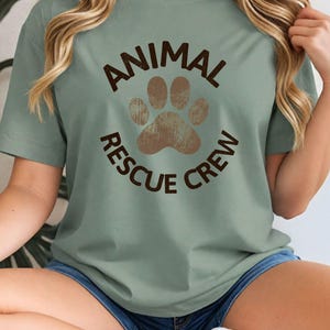 May include: Sage green t-shirt with the text "ANIMAL RESCUE CREW" in brown, encircling a brown paw print. The shirt has short sleeves and a crew neck. The model is wearing denim shorts.