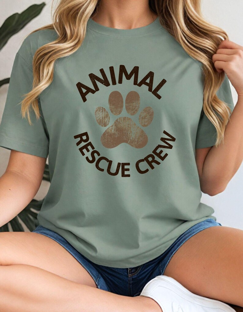 May include: Sage green t-shirt with the text "ANIMAL RESCUE CREW" in brown, encircling a brown paw print. The shirt has short sleeves and a crew neck. The model is wearing denim shorts.