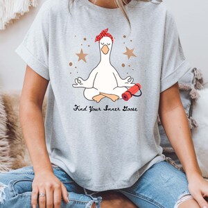 May include: A light grey t-shirt with a cartoon goose in a yoga pose, wearing a red bandana. The phrase "Find Your Inner Goose" is printed below the goose. Two brown stars and dots are also in the design.