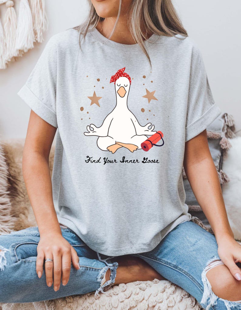 May include: A light grey t-shirt with a cartoon goose in a yoga pose, wearing a red bandana. The phrase "Find Your Inner Goose" is printed below the goose. Two brown stars and dots are also in the design.