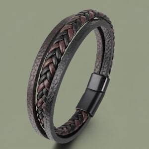 May include: A multi-strand bracelet featuring a central braided cord of black and dark brown leather, flanked by two solid black leather strips. The bracelet is secured with a smooth, rectangular black metal magnetic clasp closure.