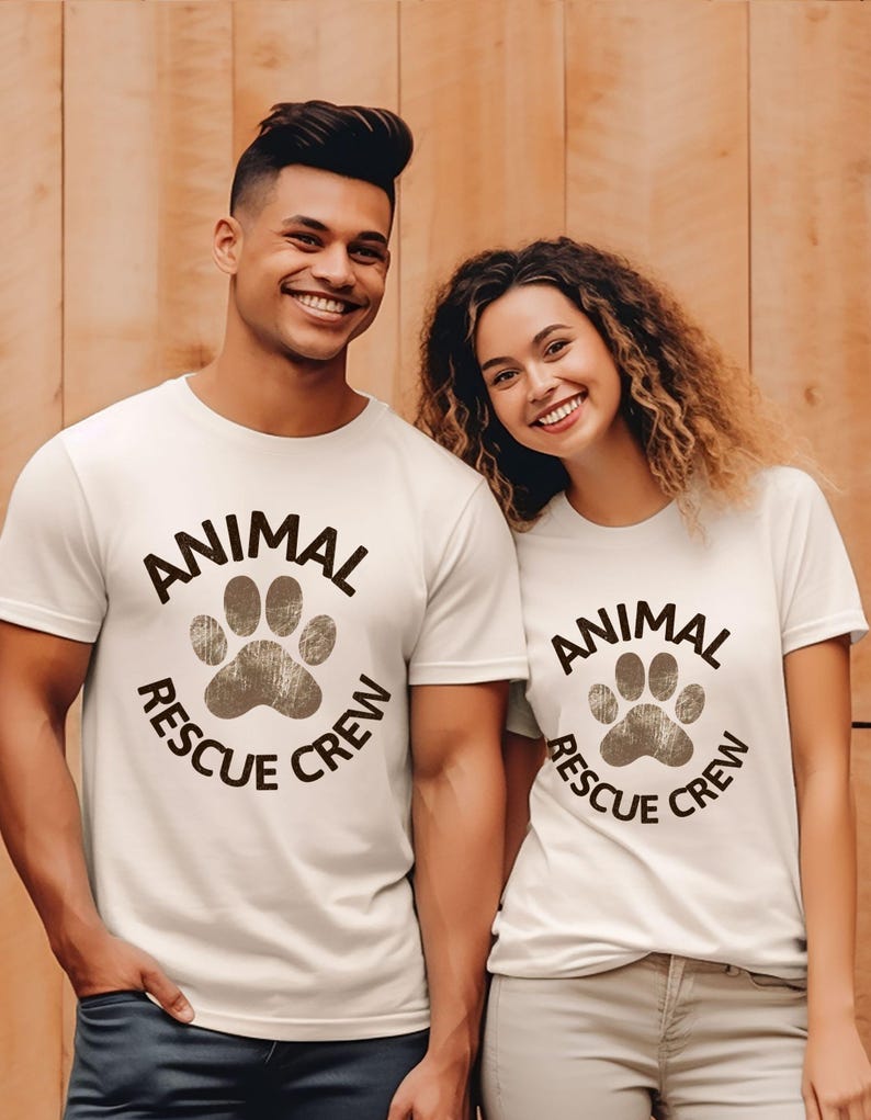 May include: Two people wearing white t-shirts with the text "ANIMAL RESCUE CREW" in a circular design with a paw print. The shirts are a light color, and the text is dark brown.