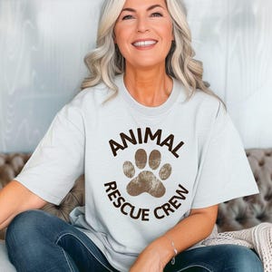 May include: A light blue t-shirt with the words "ANIMAL RESCUE CREW" encircling a brown paw print. The shirt is worn by a person with gray hair and blue jeans.