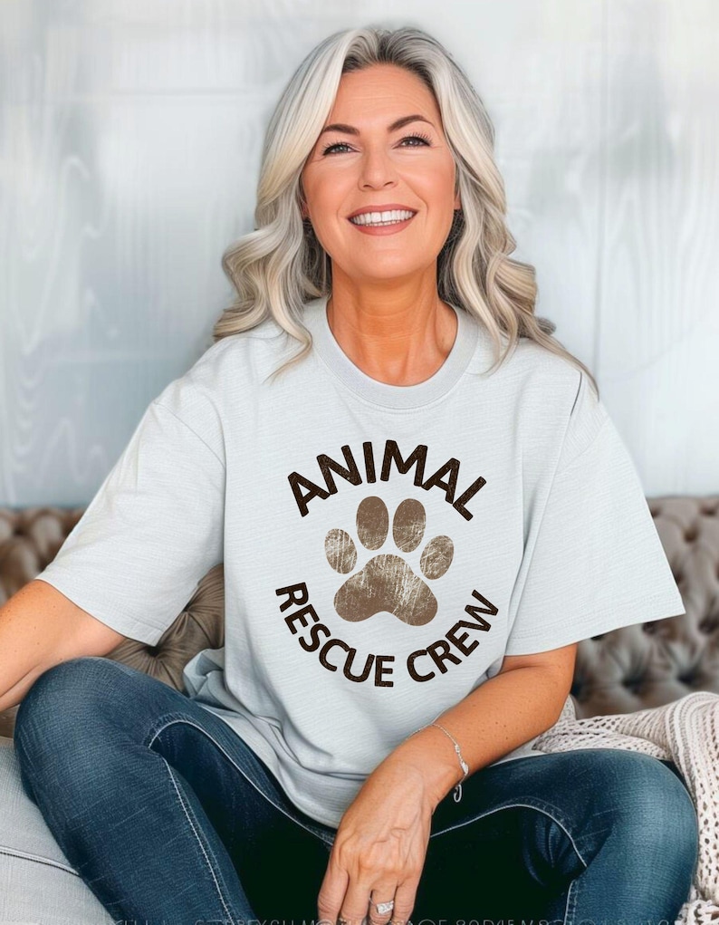 May include: A light blue t-shirt with the words "ANIMAL RESCUE CREW" encircling a brown paw print. The shirt is worn by a person with gray hair and blue jeans.