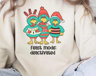Può includere: Una felpa girocollo beige con una grafica raffigurante tre tacchini color verde acqua che indossano maglioni e cappelli natalizi colorati. I tacchini presentano dettagli rossi e verdi. Sotto la grafica, il testo recita "Feast mode: deactivated" in un carattere nero.