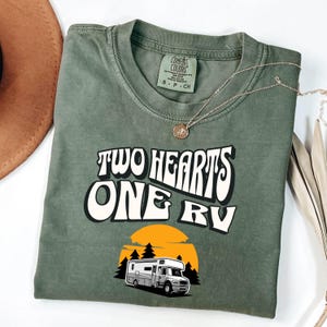Pu&ograve; includere: T-shirt verde oliva con la scritta "TWO HEARTS ONE RV" in un font retr&ograve;. Sotto, una grafica di un camper davanti a un tramonto e alberi. Una collana d'oro &egrave; drappeggiata sulla maglietta.