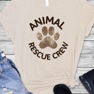 May include: Beige t-shirt with the words "ANIMAL RESCUE CREW" in brown, encircling a brown paw print. The shirt is paired with blue jeans and black shoes on a light wood background.