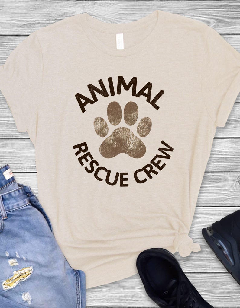 May include: Beige t-shirt with the words "ANIMAL RESCUE CREW" in brown, encircling a brown paw print. The shirt is paired with blue jeans and black shoes on a light wood background.