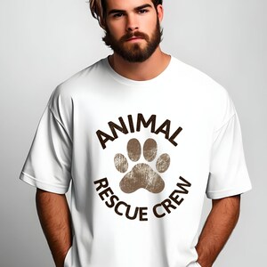 May include: White t-shirt with a brown paw print and the words "ANIMAL RESCUE CREW" in a circular design. The shirt has short sleeves and a relaxed fit.