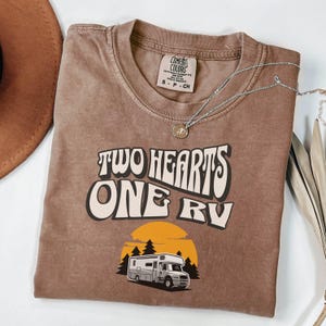 Pu&ograve; includere: T-shirt marrone con la scritta "TWO HEARTS ONE RV" in un font retr&ograve;. Il design include una grafica di un camper davanti a un tramonto e alberi. Una collana d'argento con un ciondolo &egrave; drappeggiata sulla maglietta.