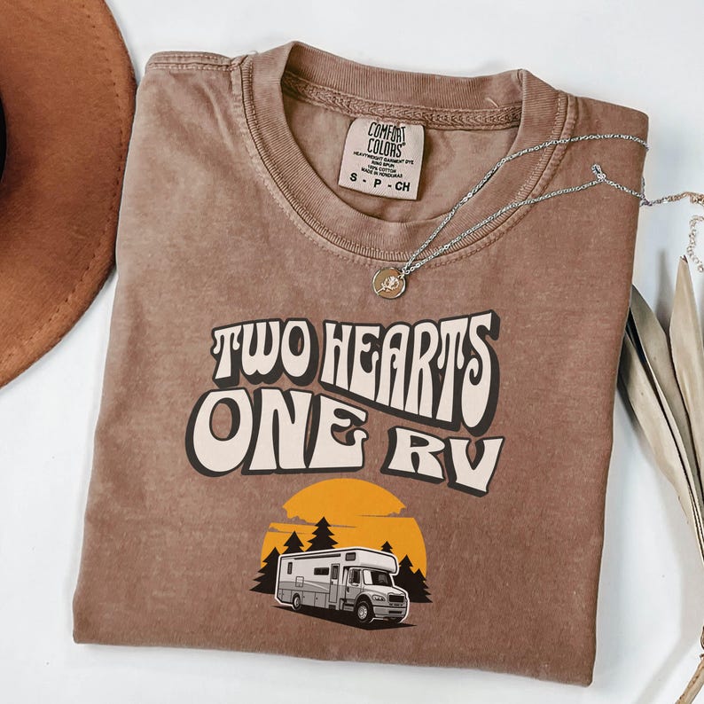 Pu&ograve; includere: T-shirt marrone con la scritta "TWO HEARTS ONE RV" in un font retr&ograve;. Il design include una grafica di un camper davanti a un tramonto e alberi. Una collana d'argento con un ciondolo &egrave; drappeggiata sulla maglietta.