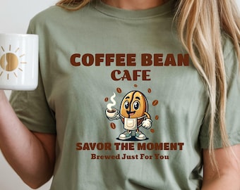 May include: Sage green t-shirt with the text "COFFEE BEAN CAFE" in brown. A cartoon coffee bean character holding a cup of coffee is featured below the text. The phrase "SAVOR THE MOMENT Brewed Just For You" is printed below the character.