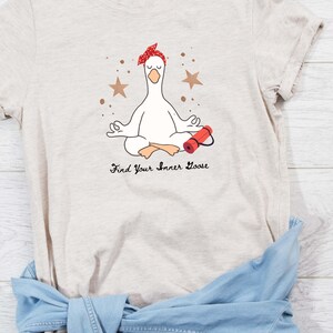 May include: Beige t-shirt with a cartoon goose meditating, wearing a red headband and with a yoga mat. The text "Find Your Inner Goose" is printed below. Brown star and dot accents surround the goose.