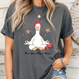 May include: Dark grey t-shirt with a cartoon goose in a yoga pose, wearing a red bandana. The shirt has the text "Find Your Inner Goose" below the goose, with gold stars and a red yoga mat graphic.