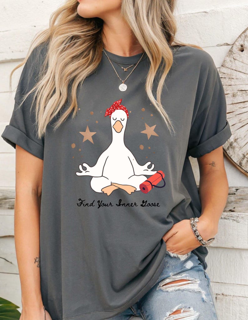 May include: Dark grey t-shirt with a cartoon goose in a yoga pose, wearing a red bandana. The shirt has the text "Find Your Inner Goose" below the goose, with gold stars and a red yoga mat graphic.