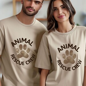 May include: Two people wearing matching beige t-shirts. Each shirt features a brown paw print design with the words "ANIMAL RESCUE CREW" in a circular arrangement. The shirts have short sleeves and a crew neck.