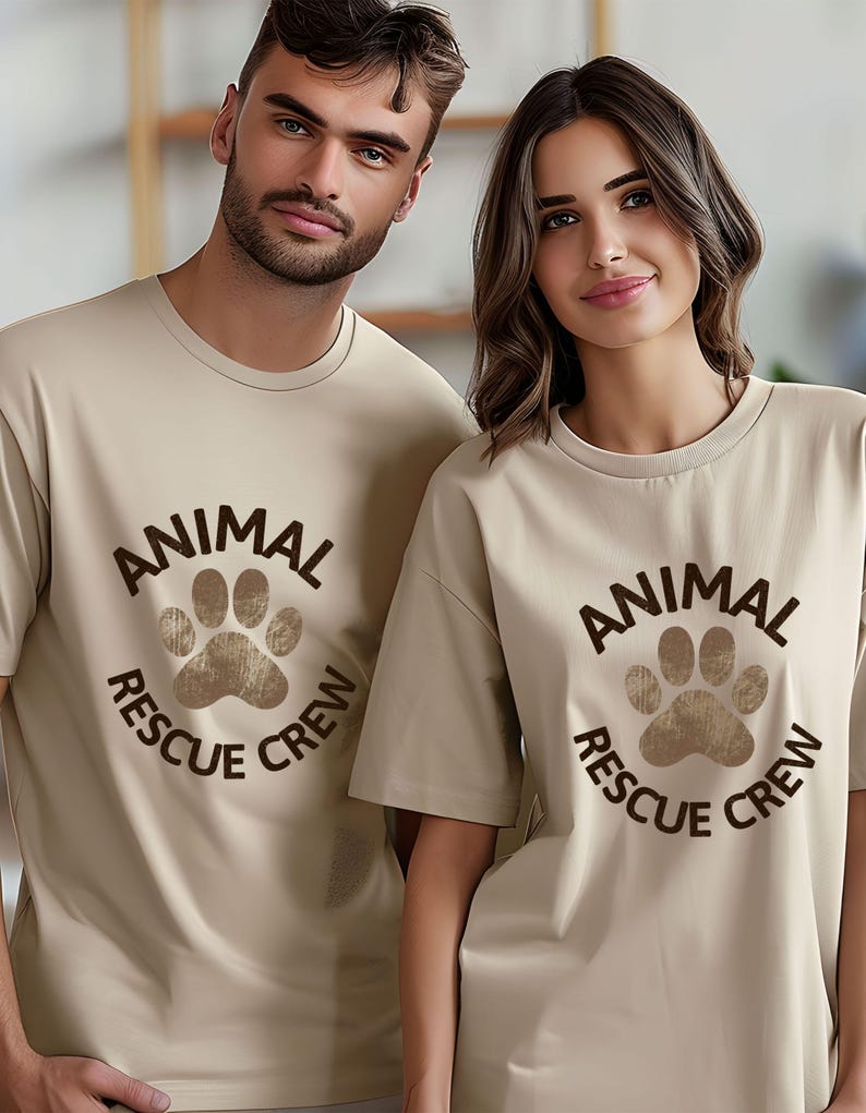 May include: Two people wearing matching beige t-shirts. Each shirt features a brown paw print design with the words "ANIMAL RESCUE CREW" in a circular arrangement. The shirts have short sleeves and a crew neck.