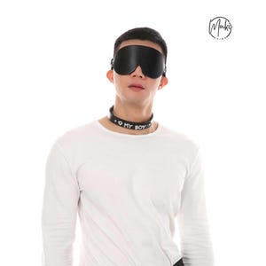 Op de afbeelding: Een zwart lederen blinddoek en een zwarte lederen halsband met een zilverkleurig hartpictogram en de tekst MY BOY in witte letters. De halsband is bevestigd met een metalen gesp en de blinddoek wordt op zijn plaats gehouden door een verstelbare band.