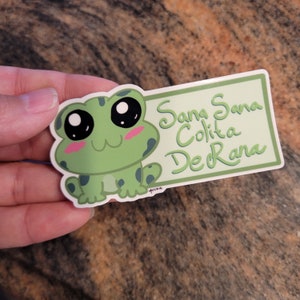 May include: A green frog sticker with big eyes and a pink blush. The sticker has a white background with the text "Sana Sana Colita De Rana" in green.