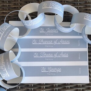 May include: A blue and white paper chain with the names of saints and the phrase "Pray For Us" printed on each link. The saints include St. Monica, St. Augustine, St. Ignatius, St. Kateri, St. Joseph, St. Francis of Assisi, St. Teresa of Avila, St. George, and St. Thomas.