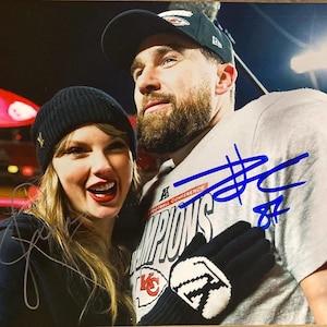 Puede incluir: Fotografía de dos personas sonriendo. Una persona lleva un gorro negro con un emblema dorado y una chaqueta negra. La otra persona lleva una camiseta gris con las palabras "FOOTBALL CONFERENCE CHAMPIONS" y una gorra negra. Ambos están sonriendo.