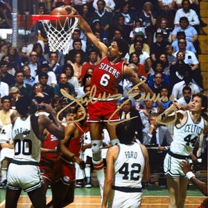 May include: A vintage photograph captures a basketball game. A player in a red Sixers jersey, number 6, dunks the ball. Other players in white and green jerseys are visible. The crowd fills the background.