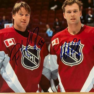 May include: Two men wearing red and white hockey jerseys with the NHL logo and a Canadian flag patch. The jerseys are signed in blue ink. The background shows a hockey arena.