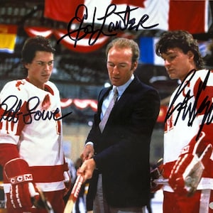 May include: A signed photograph depicting three men, possibly actors, in a hockey-themed scene. Two are wearing hockey jerseys with red and white stripes, and the third is in a suit. The image includes signatures, suggesting it's a collectible item.