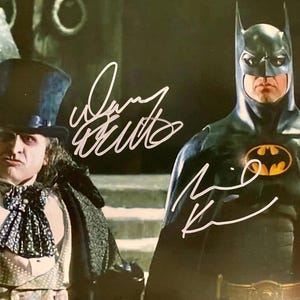 Może przedstawiać: Czarny i szary kostium Batmana z żółtym symbolem nietoperza na piersi. Mężczyzna w czarnym cylindrze i czarnym płaszczu stoi obok Batmana. Obraz jest podpisany przez Batmana i Pingwina.