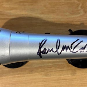 Könnte beinhalten: Silbernes Mikrofon mit schwarzer Unterschrift "Paul McCartney". Das Mikrofon ruht auf zwei schwarzen, kreisförmigen Ständern. Es hat einen Mesh-Grill und ein geripptes Design.