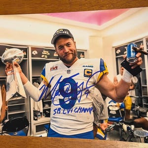 Puede incluir: Fotografía firmada de un hombre con una camiseta de fútbol blanca y azul, sosteniendo un trofeo y una lata de cerveza. La camiseta tiene el número 9 y el texto "SB LVI CHAMP". El hombre lleva una gorra negra.