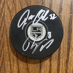 Può includere: Disco da hockey nero con il logo dei Los Angeles Kings al centro. Il disco presenta firme bianche e i numeri "32" e "8". Il disco è su una superficie di legno.