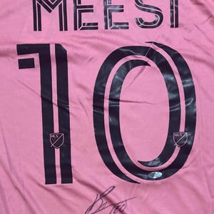 Peut inclure: Maillot de football rose avec le nom "MEESI" et le numéro "10" en noir. Comprend le logo MLS et une signature. Un maillot d'équipe sportive.