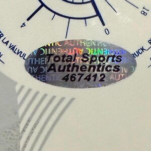 Puede incluir: Primer plano de un objeto blanco con marcas azules y el texto "Total Sports Authentics" en una pegatina hologr&aacute;fica. La pegatina tambi&eacute;n tiene el n&uacute;mero "467412".