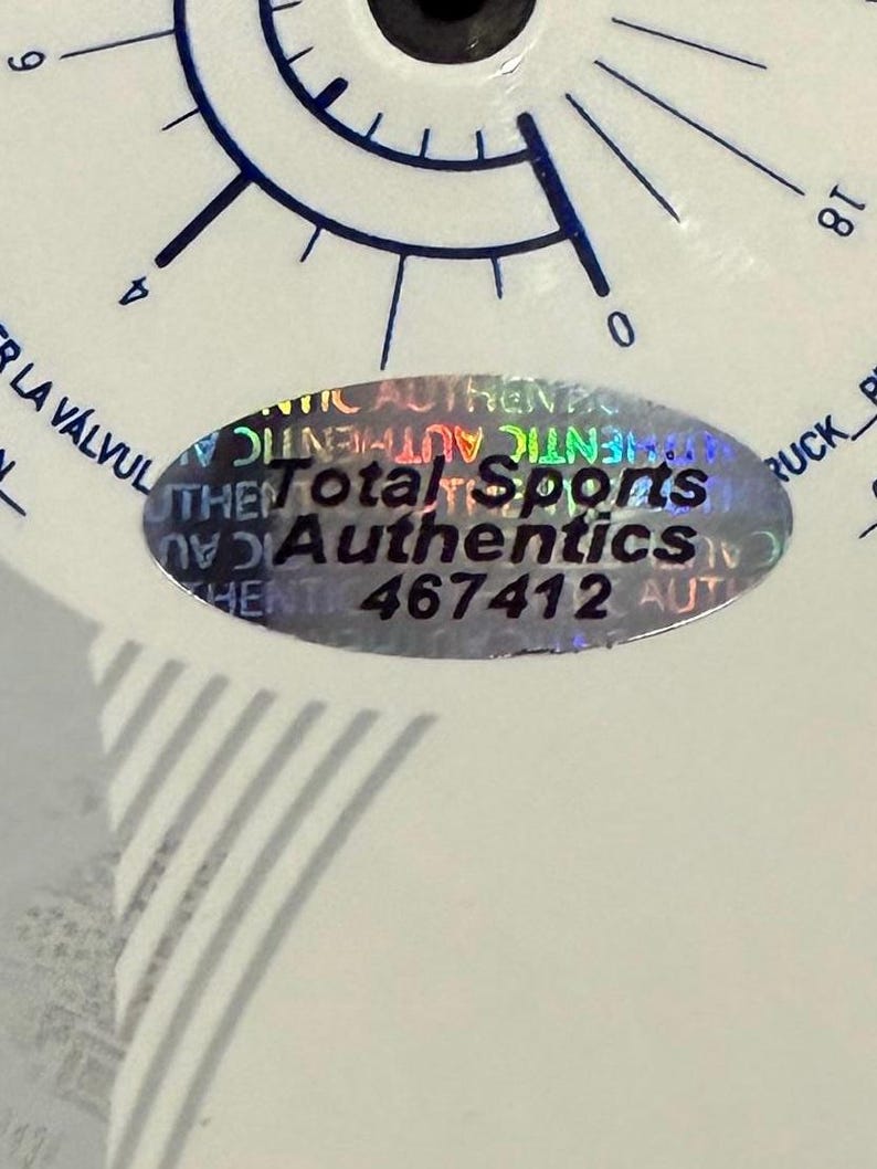 Puede incluir: Primer plano de un objeto blanco con marcas azules y el texto "Total Sports Authentics" en una pegatina hologr&aacute;fica. La pegatina tambi&eacute;n tiene el n&uacute;mero "467412".